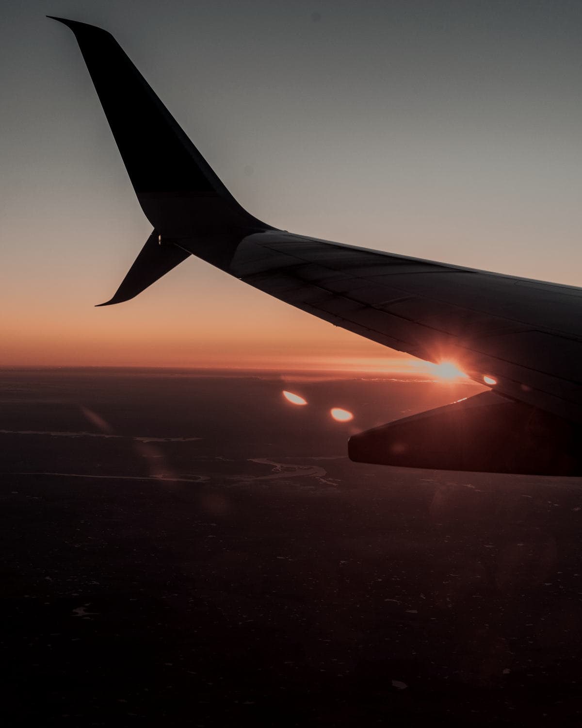 An airliner's wing above the land at dusk, the sun burning on the horizon below it