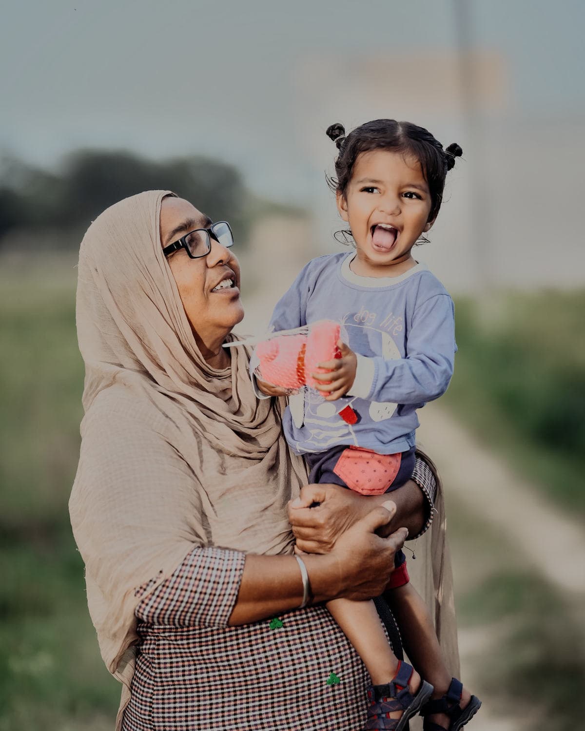 A woman in a pale orna and glasses laughs as she carries a small child on her hip on a sunlit lane