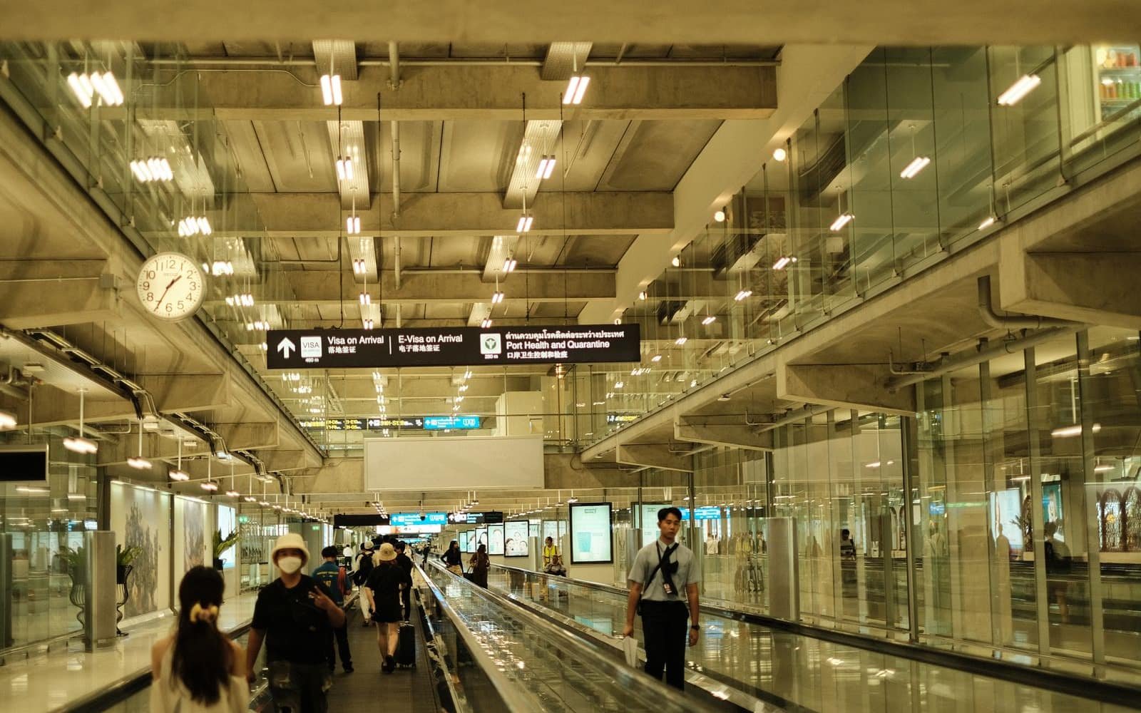 A bright, modern airport concourse lined with glass walls and a long moving walkway. A few travellers move through beneath overhead signs pointing to Visa on Arrival and Port Health, a round clock on the left wall, warm daylight filling the space.