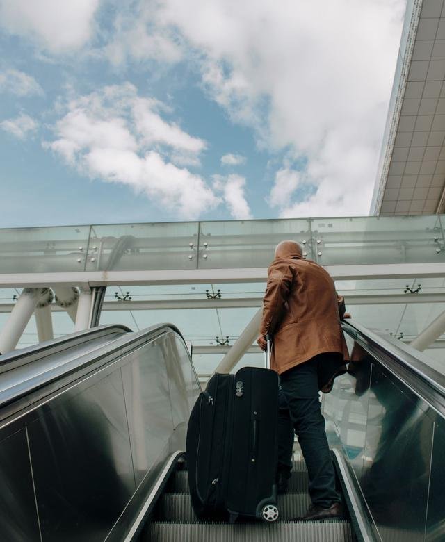 A traveller rides an airport escalator upward with a black wheeled suitcase beside him, a glass roof and open sky above.