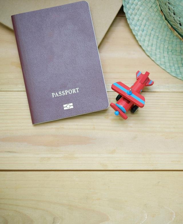 An unmarked passport, a woven sun hat and a small toy aeroplane laid out together on pale wood.