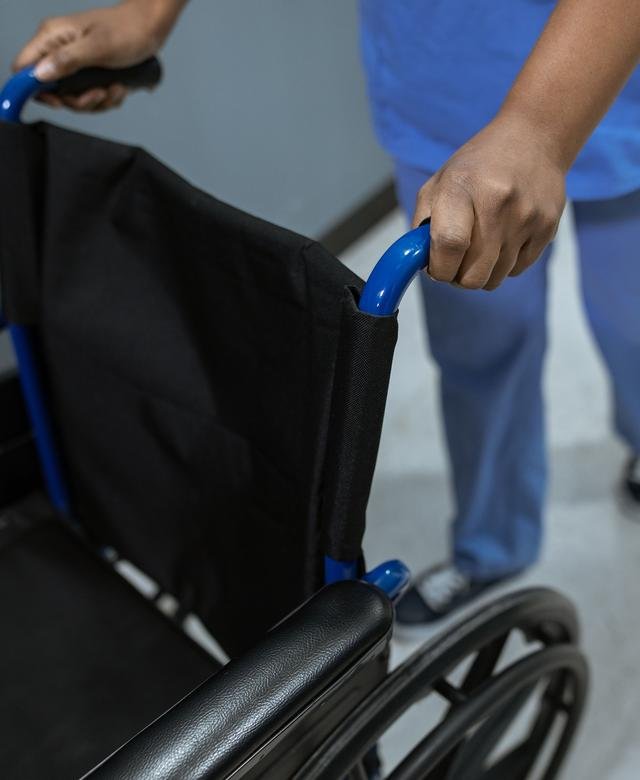 An attendant in blue scrubs wheels an empty folding wheelchair along a bright corridor, one hand on the handle.