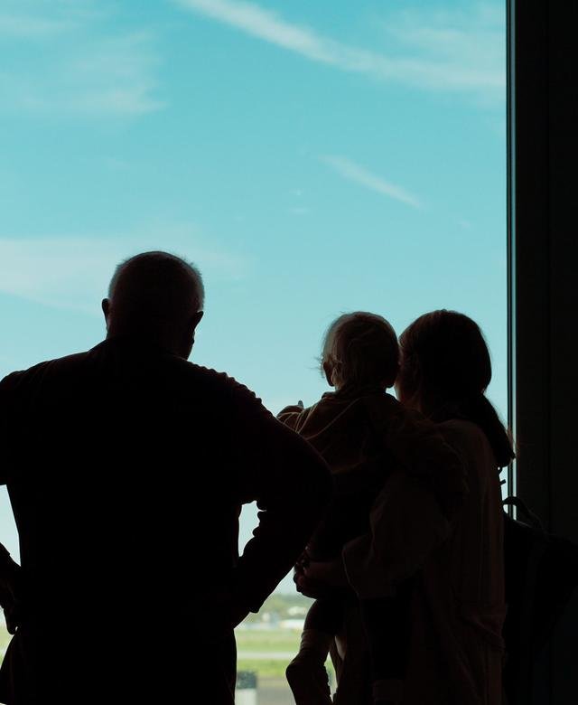 The silhouettes of a family standing at a tall airport window against a bright turquoise sky, one figure apart from the group.