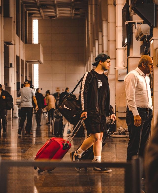 A traveller wheels a red suitcase through a warm, daylit terminal hall while other passengers wait with their bags along the wall.