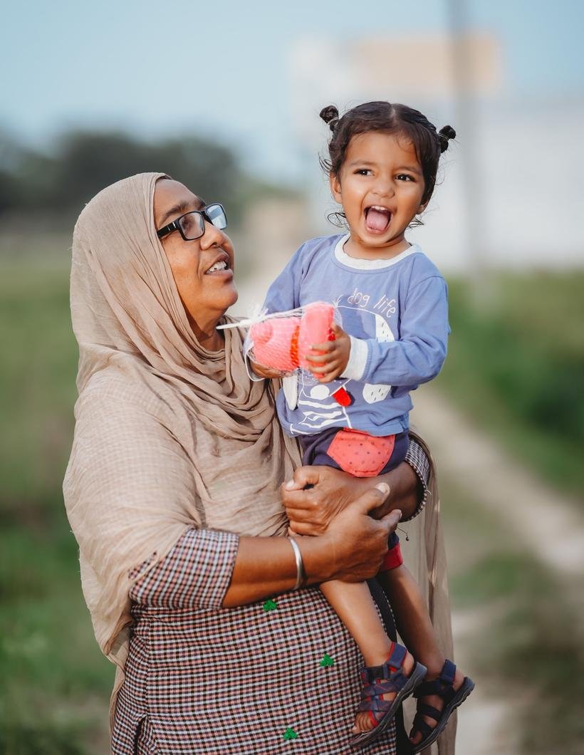 A Bangladeshi woman in a pale orna and glasses laughs as she carries a small child on her hip on a sunlit lane, the child open-mouthed with delight.