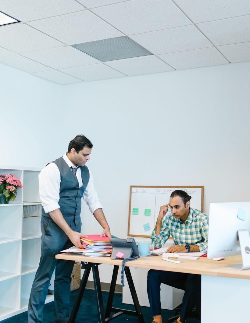 Two South Asian colleagues work together in a bright office, one standing over a desk sorting papers while the other looks up from a computer.