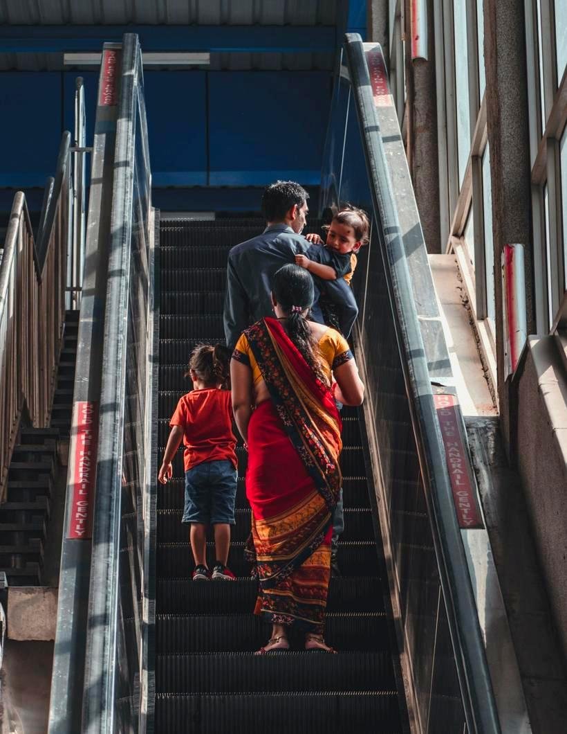 A South Asian family riding an escalator up together in a bright steel-and-glass transit building, a father carrying a toddler, a mother in a red and gold sari behind him, and a small child climbing a step ahead.