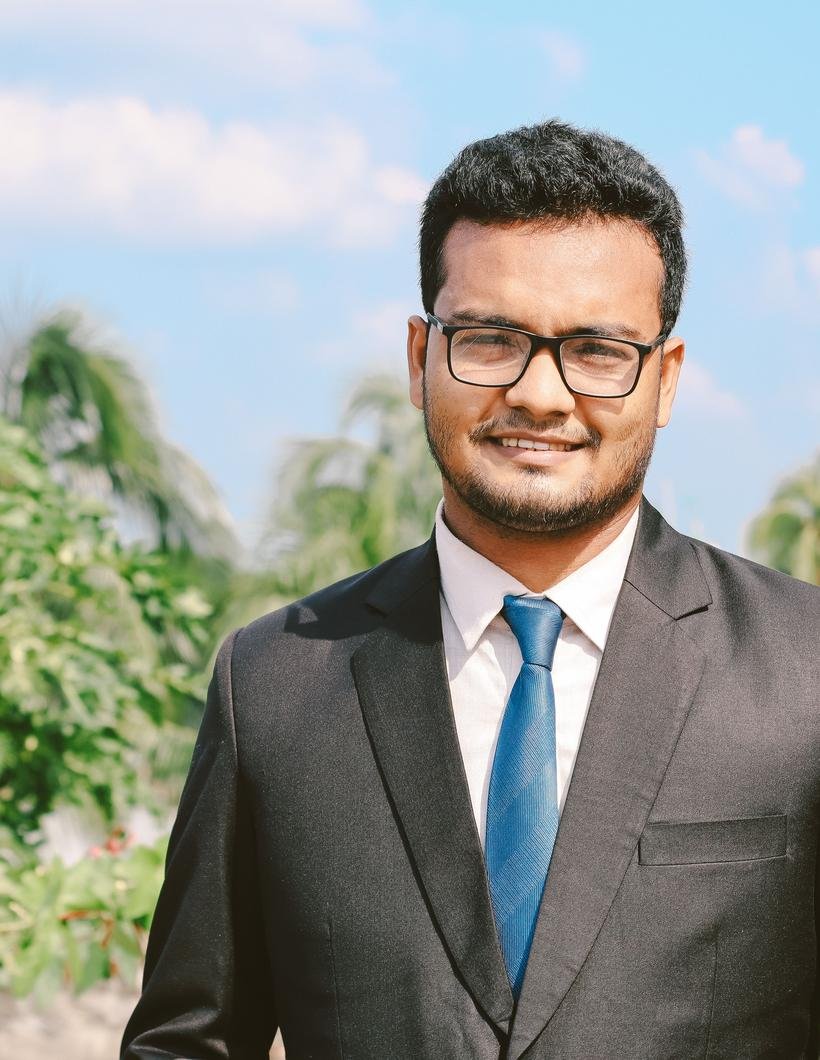 A South Asian businessman in a charcoal suit, blue tie and glasses smiles at the camera, palms and bright sky behind him.