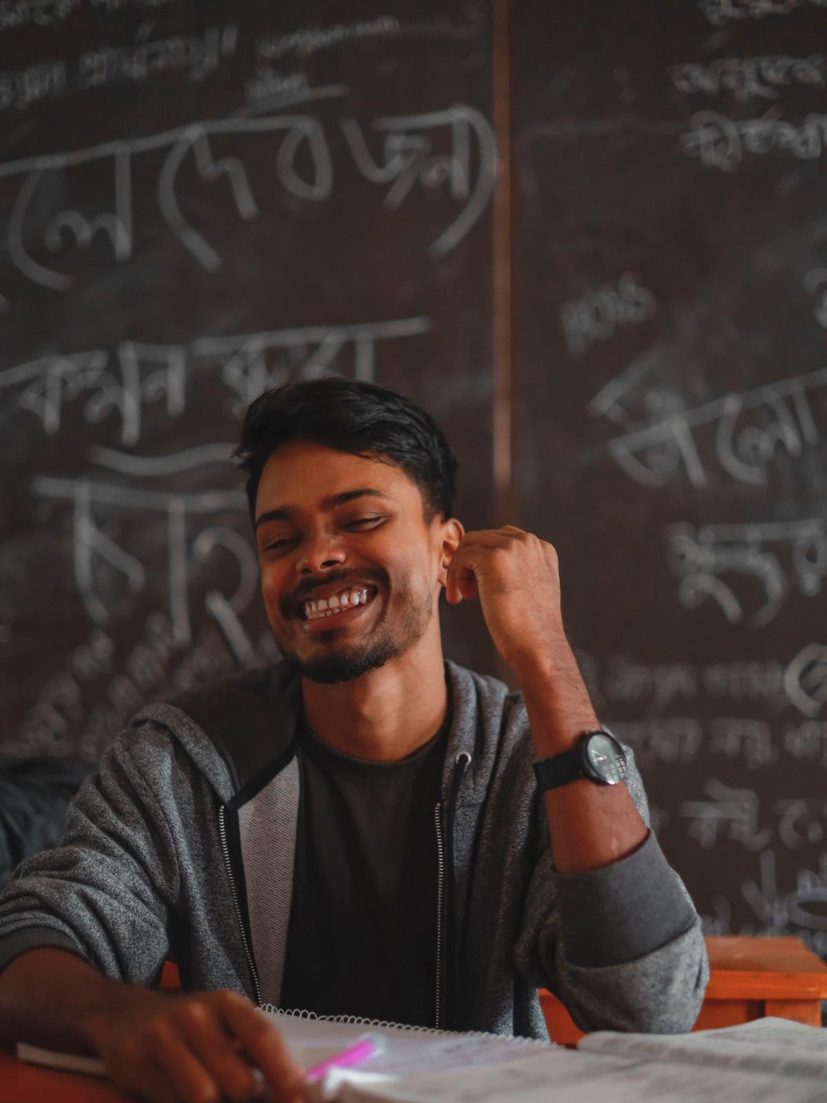A young Bangladeshi student laughing at his desk over an open notebook, a blackboard covered in Bangla writing behind him