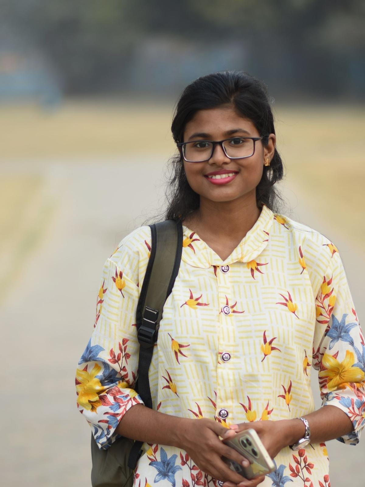 A Bangladeshi student in a printed salwar kameez and glasses, a backpack over one shoulder, smiling on a wide campus field in winter haze
