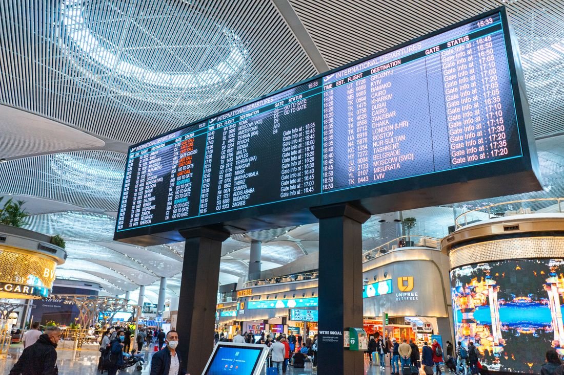 An airport departure board above a hall of passing travellers