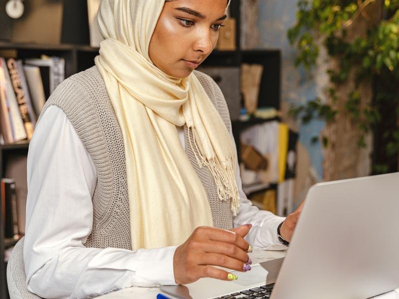 A young South Asian woman in a cream headscarf working at a laptop, printed pages spread on the desk beside her