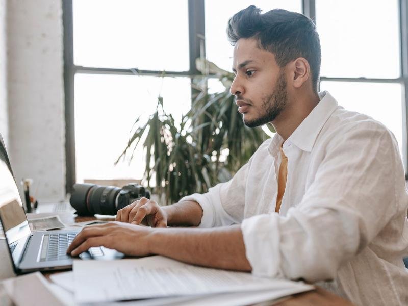 A South Asian man in a white shirt working at a laptop with papers spread beside him