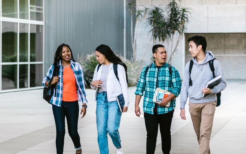 Four students with backpacks and books walking together across a campus plaza